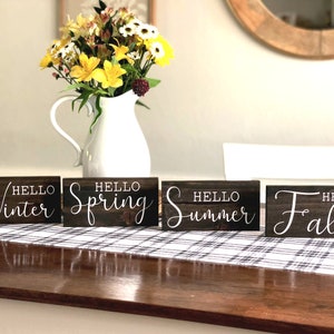 May include: Four wooden signs with the words "Hello Winter", "Hello Spring", "Hello Summer", and "Hello Fall" written in white cursive font. The signs are arranged in a row on a wooden table with a black and white checkered tablecloth.