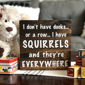 May include: A wooden sign with the text "I don't have ducks... or a row... I have SQUIRRELS and they're EVERYWHERE". The sign is brown with white lettering and is sitting on a table with a stuffed animal and wooden blocks.