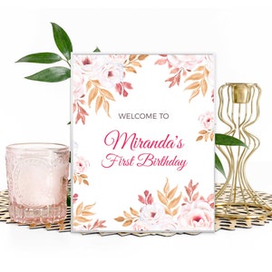 May include: A white welcome sign with a floral design in shades of pink, brown, and gold. The text on the sign reads "Welcome to Miranda's First Birthday".
