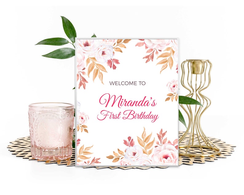 May include: A white welcome sign with a floral design in shades of pink, brown, and gold. The text on the sign reads "Welcome to Miranda's First Birthday".
