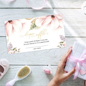 Puede incluir: Una tarjeta de rifa de pa&ntilde;ales con dise&ntilde;o floral rosa y blanco y texto dorado que dice "diaper raffle" y "bring a pack of diaper, in any size, for your chance to win a special prize!" con una l&iacute;nea en blanco para "your name".