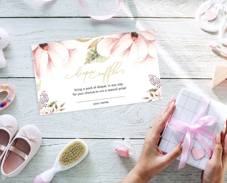 Puede incluir: Una tarjeta de rifa de pa&ntilde;ales con dise&ntilde;o floral rosa y blanco y texto dorado que dice "diaper raffle" y "bring a pack of diaper, in any size, for your chance to win a special prize!" con una l&iacute;nea en blanco para "your name".