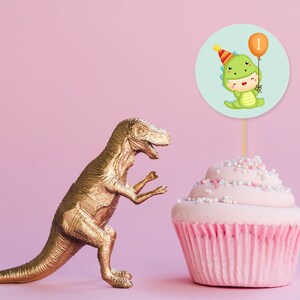May include: A pink cupcake with colorful sprinkles topped with a green dinosaur wearing a party hat and holding a balloon with the number 1 on it. A gold dinosaur figurine is in the background.