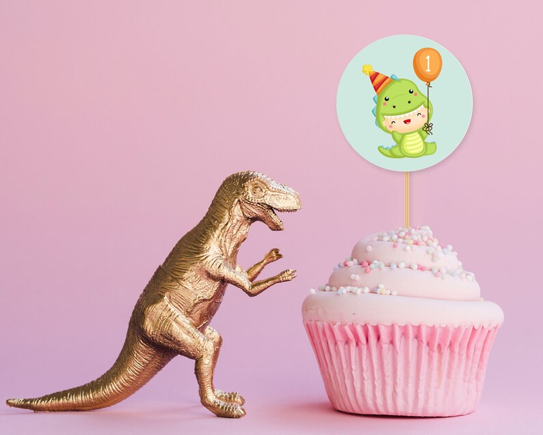 May include: A pink cupcake with colorful sprinkles topped with a green dinosaur wearing a party hat and holding a balloon with the number 1 on it. A gold dinosaur figurine is in the background.