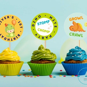 May include: Three cupcakes decorated with green, yellow, and blue frosting. Each cupcake has a round, colorful paper topper with dinosaur-themed text. The toppers read "Oliver's 1st Birthday", "Dino Party Stomp Chomp", and "Growl Crawl".