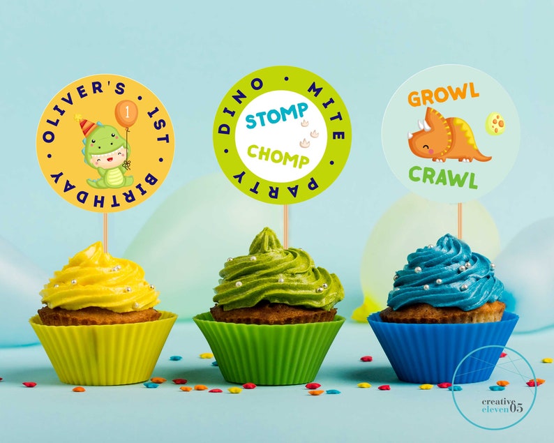 May include: Three cupcakes decorated with green, yellow, and blue frosting. Each cupcake has a round, colorful paper topper with dinosaur-themed text. The toppers read "Oliver's 1st Birthday", "Dino Party Stomp Chomp", and "Growl Crawl".