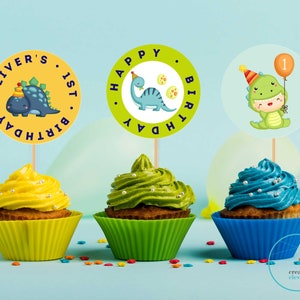 May include: Three cupcakes decorated with green, yellow, and blue frosting. Each cupcake has a round, colorful paper topper with a dinosaur design and the text "Oliver's 1st Birthday" and "Happy Birthday".