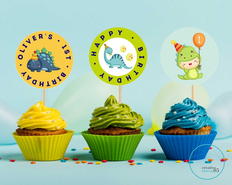 May include: Three cupcakes decorated with green, yellow, and blue frosting. Each cupcake has a round, colorful paper topper with a dinosaur design and the text "Oliver's 1st Birthday" and "Happy Birthday".