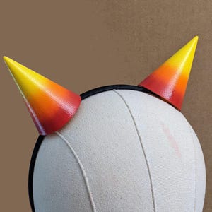 May include: A pair of costume horns with a gradient of yellow, orange, and red. The horns are attached to a black headband, displayed on a mannequin head. The horns are a fun accessory for cosplay or themed events.