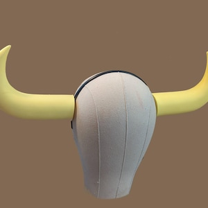 May include: A bull horn headpiece with two curved, yellow horns attached to a cream-colored head form. The horns are a bright yellow, and the head form has vertical seams. A black band is visible at the base of the horns.