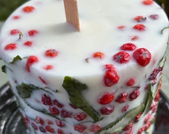 May include: A cylindrical white wax candle featuring embedded red berries and green leaves throughout the exterior and top surface. A single rectangular wooden wick is positioned in the center of the candle, extending upward from the flat top.