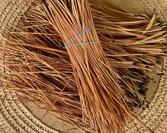 May include: A bundle of dried pine needles tied together with a blue rubber band. The needles are a light brown color and are arranged in a fan shape. The needles are sitting on a woven brown mat.