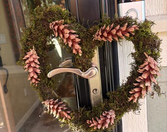 May include: A heart-shaped wreath constructed from dark green moss and decorated with several light brown pinecones. The wreath is hanging on a silver door handle attached to a black door frame with a silver smart doorbell mounted above.