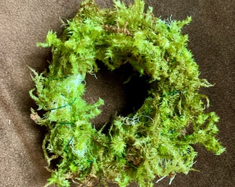 May include: A circular wreath constructed from vibrant green preserved moss. The structure features a dense, textured arrangement of natural moss fibers secured with thin, visible green wire, forming a hollow center with an irregular, organic edge.