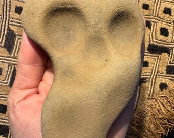 May include: A tan-colored, smooth stone carving with two eye-shaped indentations and a rounded shape. The stone is held in a hand, showcasing its texture and form. The object appears to be a unique piece of art or a decorative item.