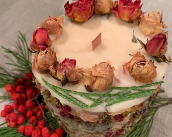 May include: A white candle with dried flowers and greenery around the base. The candle is decorated with dried roses and pine needles.