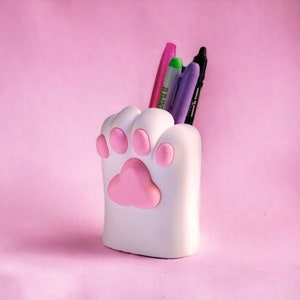 Puede incluir: Porta l&aacute;pices de cer&aacute;mica blanca con forma de pata con huellas de pata rosas. El porta l&aacute;pices tiene cuatro bol&iacute;grafos dentro, incluyendo uno rosa, uno verde, uno morado y uno negro.