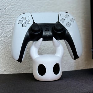 May include: A white PlayStation 5 controller is held up by a white 3D printed stand shaped like the Knight from the video game Hollow Knight.