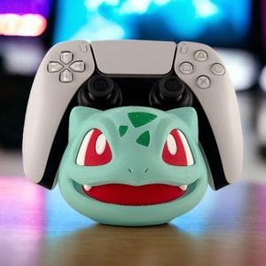 Pode incluir: Um controlador branco do PlayStation 5 com um personagem Bulbasaur Pokemon verde, vermelho e branco preso na frente.