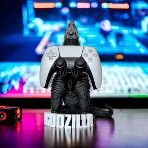 May include: A white PlayStation 5 controller is held by a black Godzilla figurine on a white base with the word "GODZILLA" printed on it.