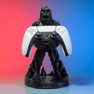 以下が含まれることがあります： 白と黒のゲームコントローラーを持つ黒いゴリラ型のコントローラーホルダー。スタンドは、テクスチャ加工された表面を持つ丸いベースにあります。背景は赤から青へのグラデーションです。