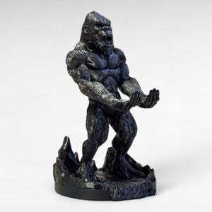 May include: A black and gray gorilla figurine in a dynamic pose, with its mouth open and arms outstretched. The gorilla is standing on a rocky base. The figurine has detailed fur and muscular definition.