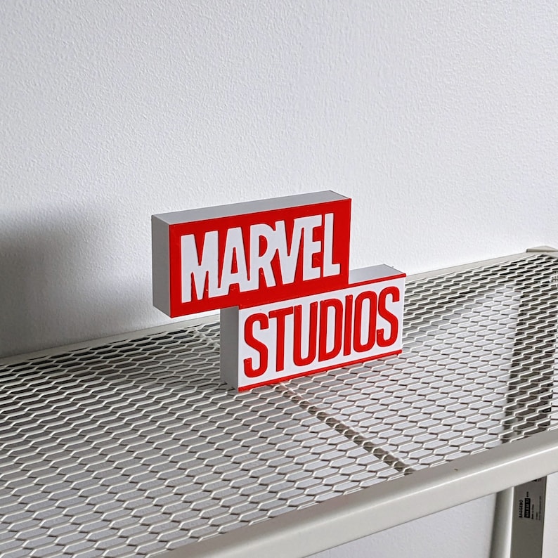 K&ouml;nnte beinhalten: Wei&szlig;es Metallregal mit einem roten und wei&szlig;en Schild mit der Aufschrift "MARVEL STUDIOS".