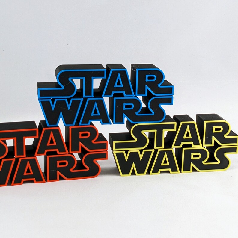 May include: Three black plastic "Star Wars" logos stacked on top of each other. The top logo is blue, the middle logo is red, and the bottom logo is yellow.