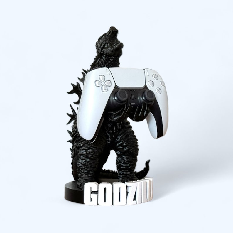 May include: A black Godzilla figure holding a white PlayStation 5 controller. The figure is standing on a base with the word "GODZILLA" printed on it.