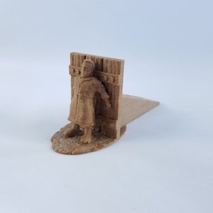 May include: A brown 3D printed doorstop shaped like a person standing in a doorway. The person is wearing a long robe and has their hands raised.