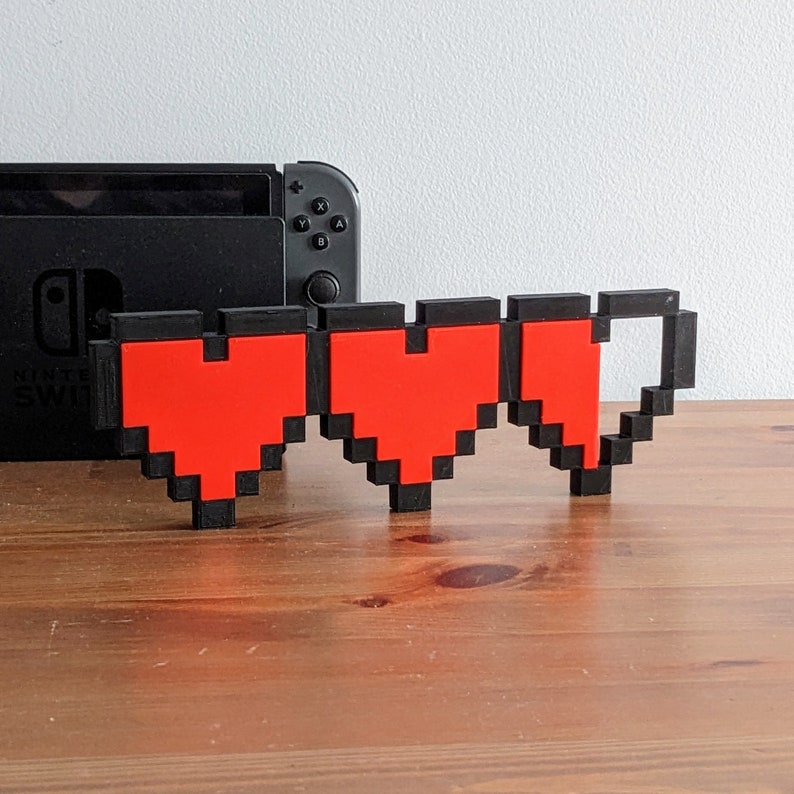 May include: Three red pixelated hearts in a row, made of black plastic, with a white outline. The hearts are arranged horizontally and are a popular video game design.
