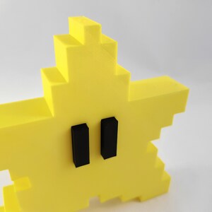 May include: A yellow pixelated star with two black rectangular eyes. The star is a 3D printed object.