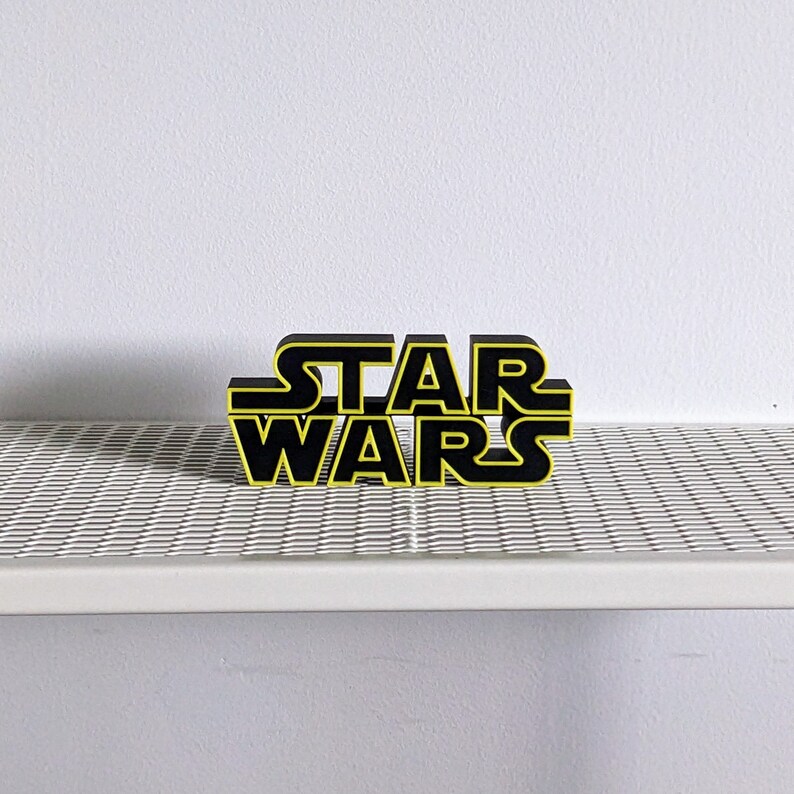 May include: Black and yellow 3D block letters spelling "STAR WARS" on a white surface.