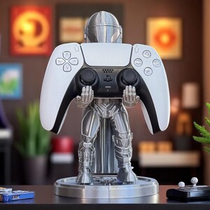 Pode incluir: Um suporte de controlador prateado com tema Mandalorian segurando um controlador PlayStation 5 branco e preto. O suporte est&aacute; em uma base redonda e apresenta um design detalhado da armadura Mandalorian. O controlador est&aacute; centralizado.