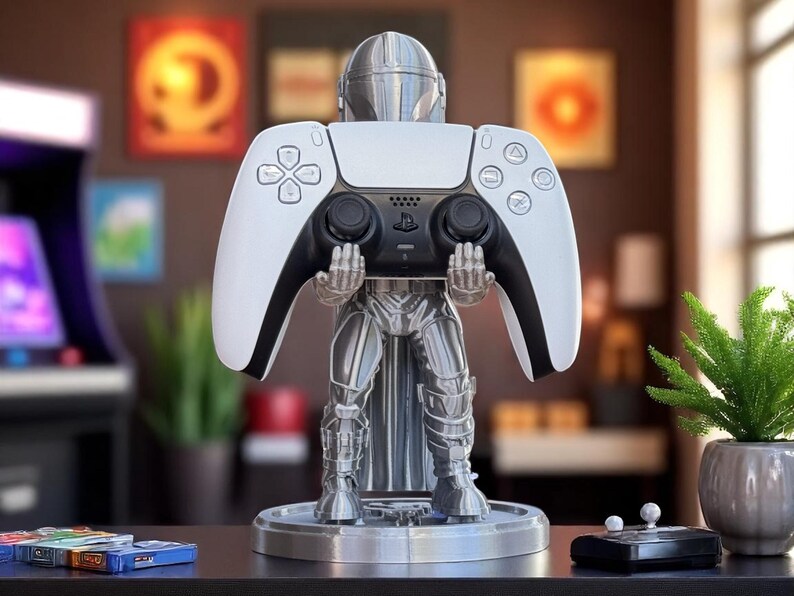 Pode incluir: Um suporte de controlador prateado com tema Mandalorian segurando um controlador PlayStation 5 branco e preto. O suporte est&aacute; em uma base redonda e apresenta um design detalhado da armadura Mandalorian. O controlador est&aacute; centralizado.