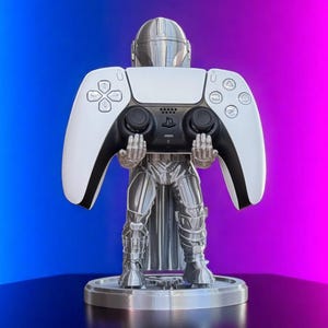 Pode incluir: Um suporte de controlador prateado com tema Mandaloriano segurando um controlador PlayStation 5 branco e preto. O suporte &eacute; redondo e tem um design detalhado da armadura do Mandaloriano. O fundo &eacute; um gradiente de azul e rosa.