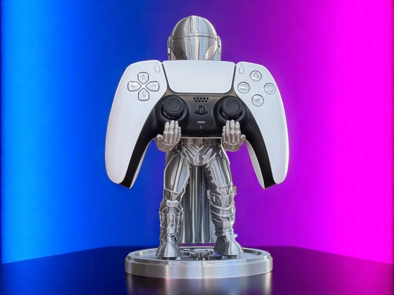 Pode incluir: Um suporte de controlador prateado com tema Mandaloriano segurando um controlador PlayStation 5 branco e preto. O suporte &eacute; redondo e tem um design detalhado da armadura do Mandaloriano. O fundo &eacute; um gradiente de azul e rosa.