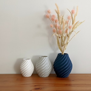 K&ouml;nnte beinhalten: Drei Keramikvasen mit einem spiralf&ouml;rmigen Muster. Die Vase rechts ist dunkelblau, die mittlere Vase ist hellgrau und die Vase links ist wei&szlig;. Die Vasen stehen auf einer Holzoberfl&auml;che.