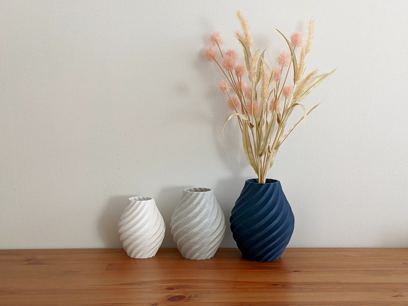 K&ouml;nnte beinhalten: Drei Keramikvasen mit einem spiralf&ouml;rmigen Muster. Die Vase rechts ist dunkelblau, die mittlere Vase ist hellgrau und die Vase links ist wei&szlig;. Die Vasen stehen auf einer Holzoberfl&auml;che.