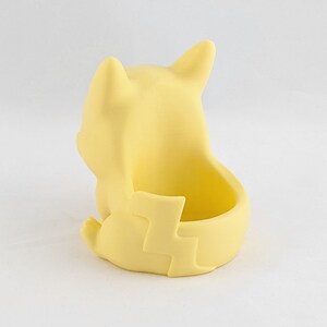 May include: A yellow ceramic planter shaped like a cartoon character sitting with its legs crossed. The planter has a small opening on the back for planting.