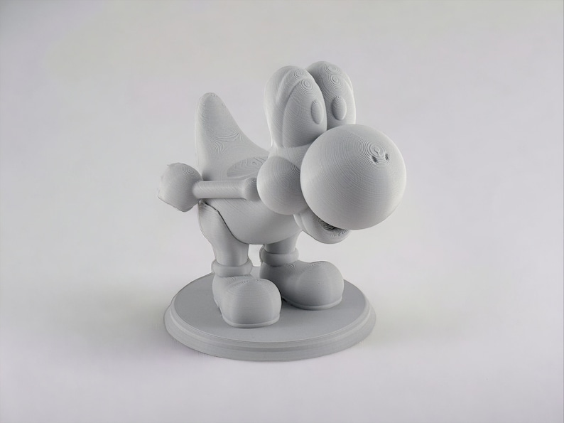 May include: A 3D printed figurine of a gray Yoshi, a dinosaur-like character from the Super Mario video game franchise. The figurine is posed in a crouching position with its head tilted down and its mouth slightly open. The figurine is standing on a circular base.