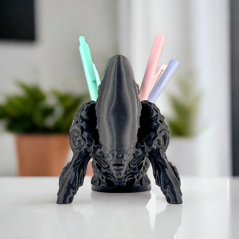 May include: Black 3D printed alien head desk organizer with three pens in it. The pens are pink, light blue, and green.