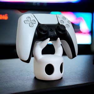 K&ouml;nnte beinhalten: Ein wei&szlig;er PlayStation 5 DualSense-Controller wird in einem wei&szlig;en 3D-gedruckten St&auml;nder gehalten, der wie der Ritter aus dem Videospiel Hollow Knight geformt ist.
