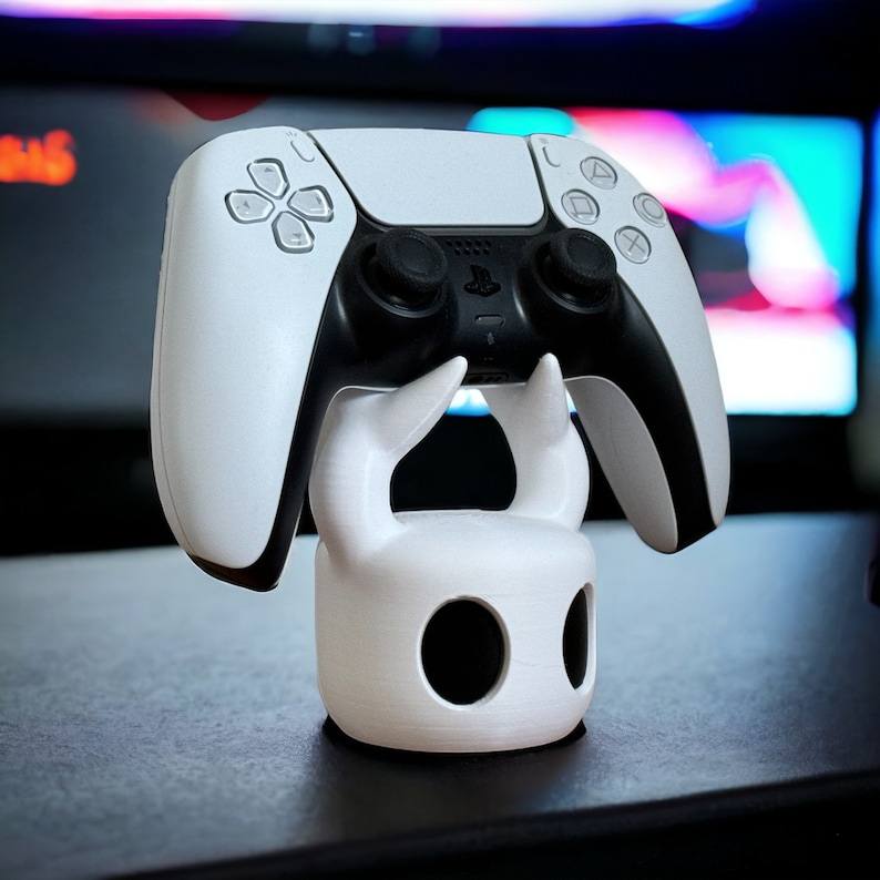 K&ouml;nnte beinhalten: Ein wei&szlig;er PlayStation 5 DualSense-Controller wird in einem wei&szlig;en 3D-gedruckten St&auml;nder gehalten, der wie der Ritter aus dem Videospiel Hollow Knight geformt ist.