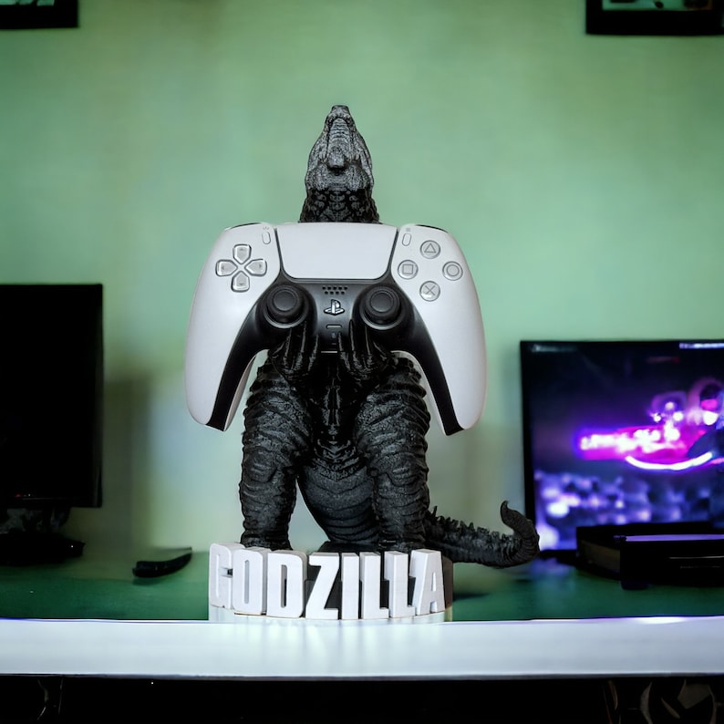 May include: A white PlayStation 5 controller is held by a black Godzilla figurine. The figurine is standing on a base with the word "GODZILLA" printed on it.