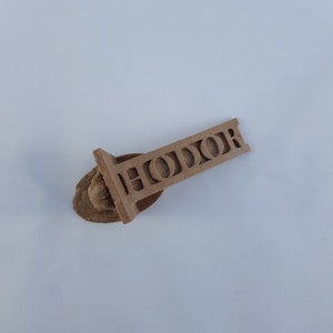 May include: A wooden keychain with the word "HODOR" carved into it. The keychain is brown and has a small, round, brown piece of wood attached to the top.