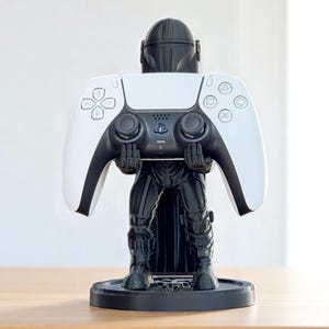 Pode incluir: Um suporte de controlador com tema Mandaloriano em preto e branco segurando um controlador PlayStation 5 branco e preto. O suporte &eacute; uma estatueta detalhada do Mandaloriano, com o controlador repousando em suas m&atilde;os. O suporte est&aacute; em uma base circular preta.