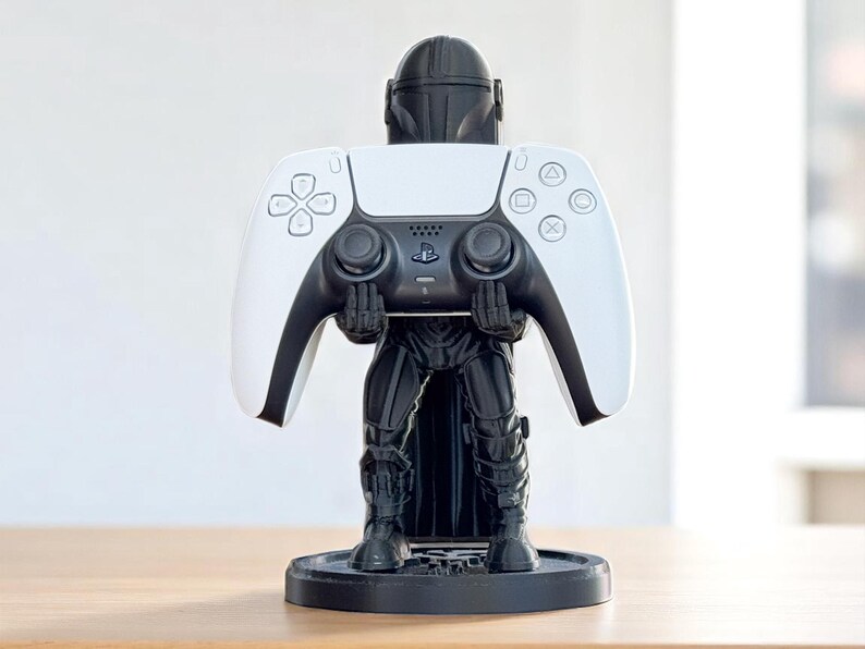 Pode incluir: Um suporte de controlador com tema Mandaloriano em preto e branco segurando um controlador PlayStation 5 branco e preto. O suporte &eacute; uma estatueta detalhada do Mandaloriano, com o controlador repousando em suas m&atilde;os. O suporte est&aacute; em uma base circular preta.