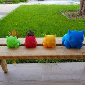 May include: Four colorful ceramic planters shaped like cartoon characters, sitting on a wooden bench. The planters are green, red, yellow, and blue.