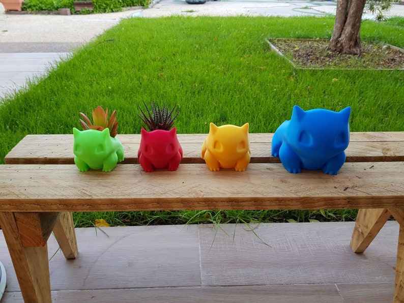 May include: Four colorful ceramic planters shaped like cartoon characters, sitting on a wooden bench. The planters are green, red, yellow, and blue.